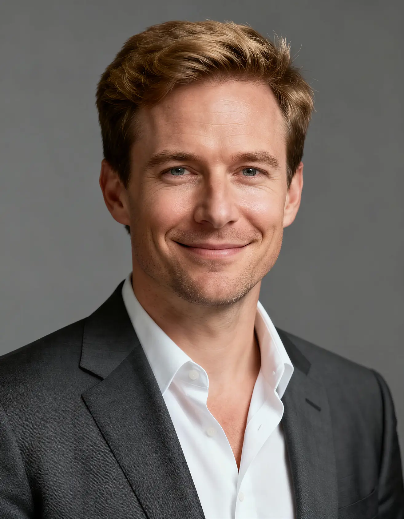 Smiling man with light brown hair wearing a dark gray suit jacket and white dress shirt against a plain gray background.