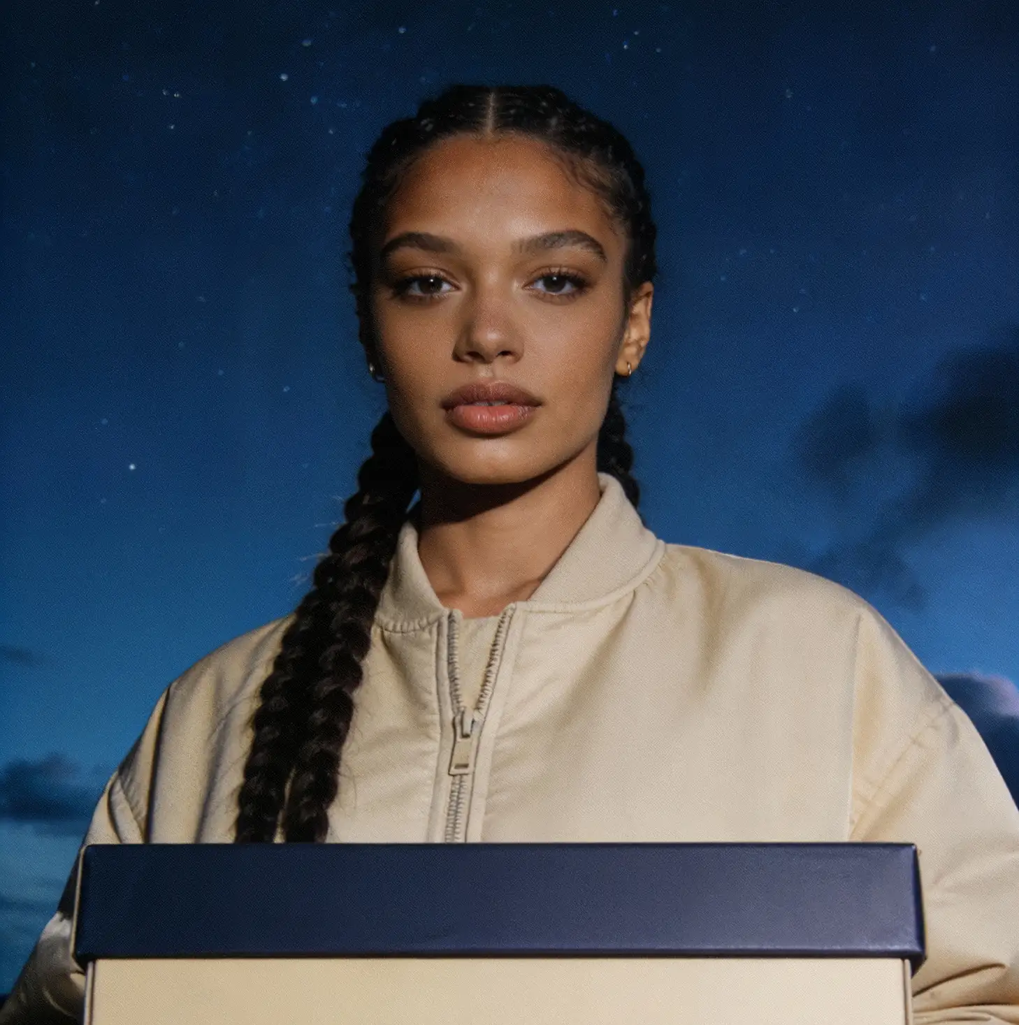 Woman with two long braids wearing a beige zip-up jacket, holding a large cream-colored box with a dark blue lid against a night sky background.