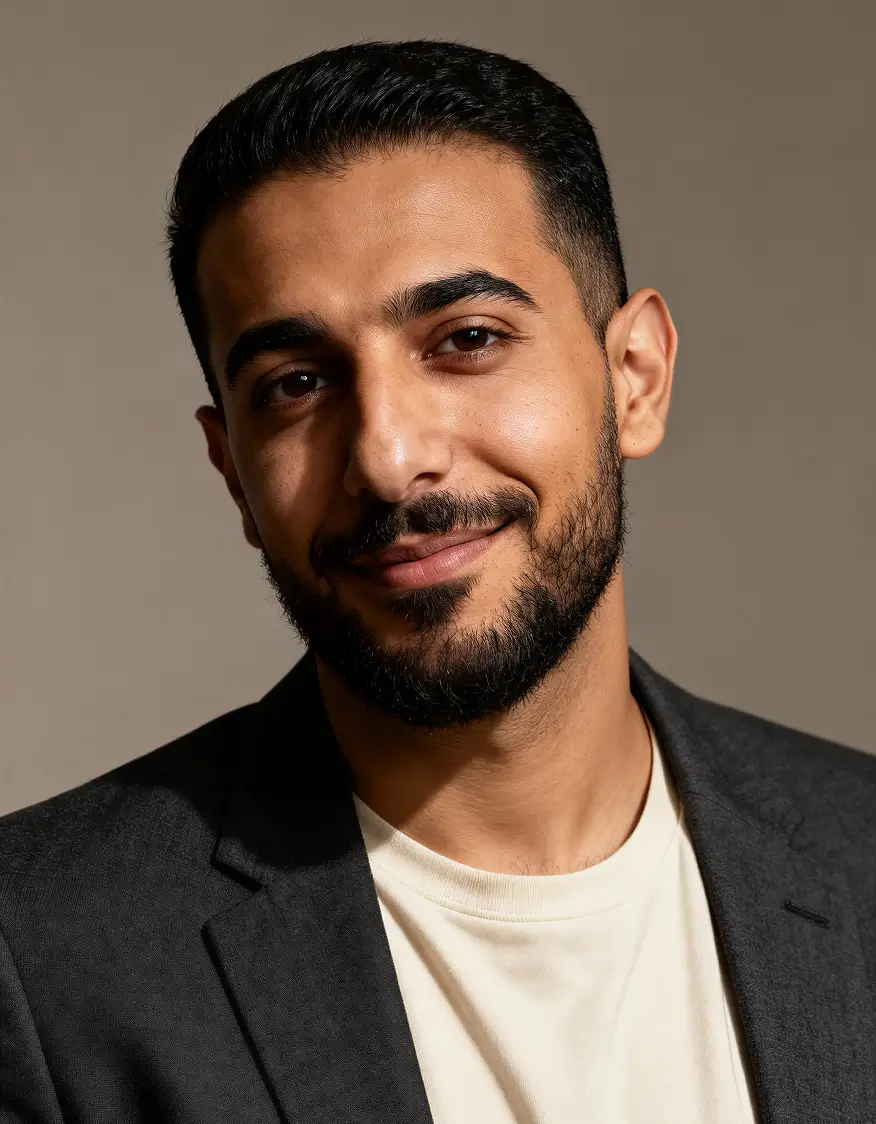 Close-up portrait of a man with short black hair and beard wearing a white shirt and dark blazer, smiling gently against a plain background.