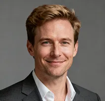Smiling man with light brown hair wearing a dark suit jacket and white shirt against a gray background.