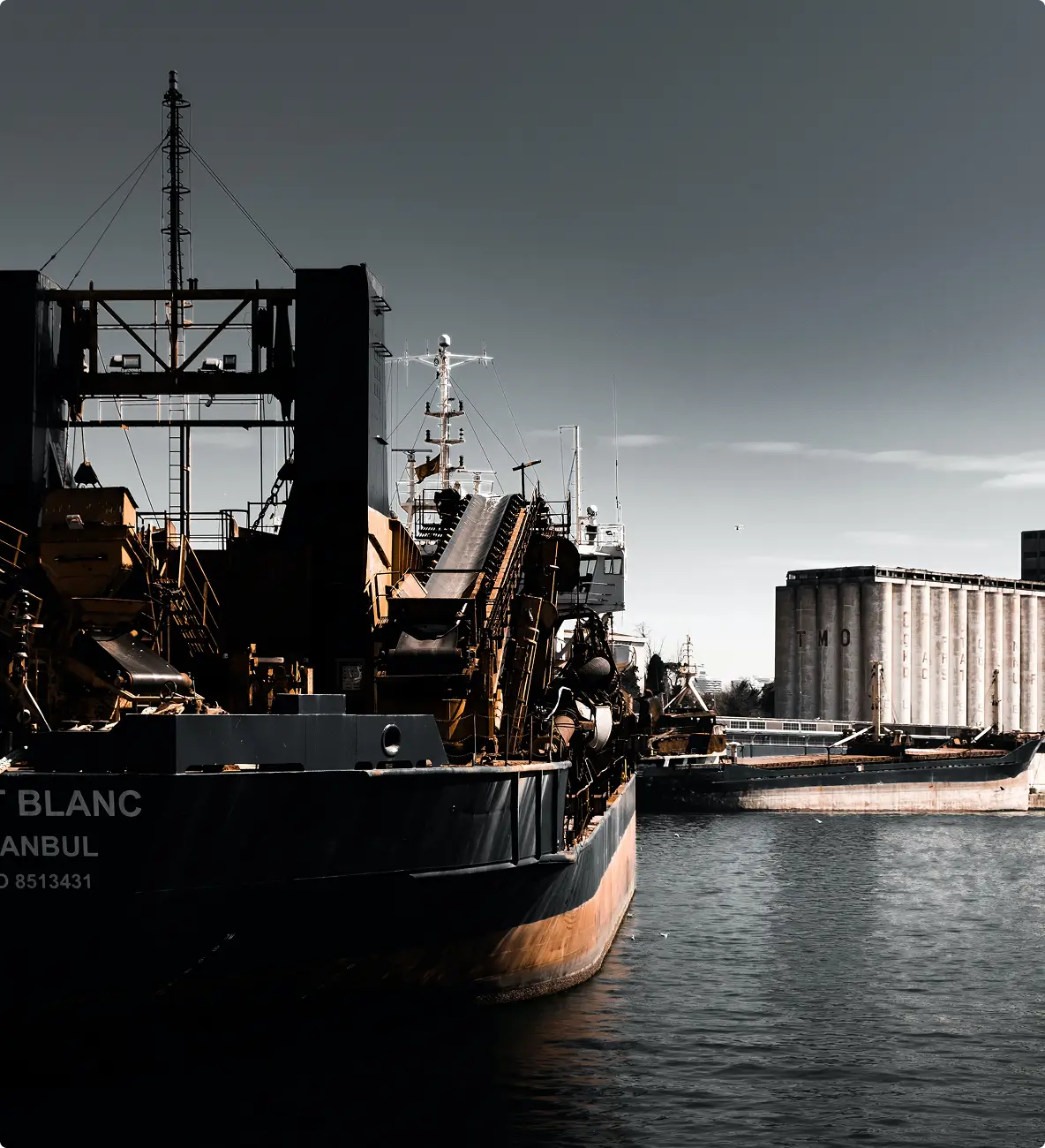 Large dark ship docked at a harbor with industrial equipment on board and a distant concrete grain elevator marked TMO.