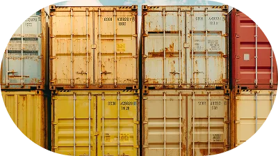 Stacked shipping containers in various faded colors including yellow, beige, and red, with some rust marks and identification codes visible.